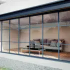 Steellook glazen schuifwand met 7 rails, vanaf buiten naar binnen kijkend, houten vloer binnen en grijze bank met gras en een paadje in de tuin